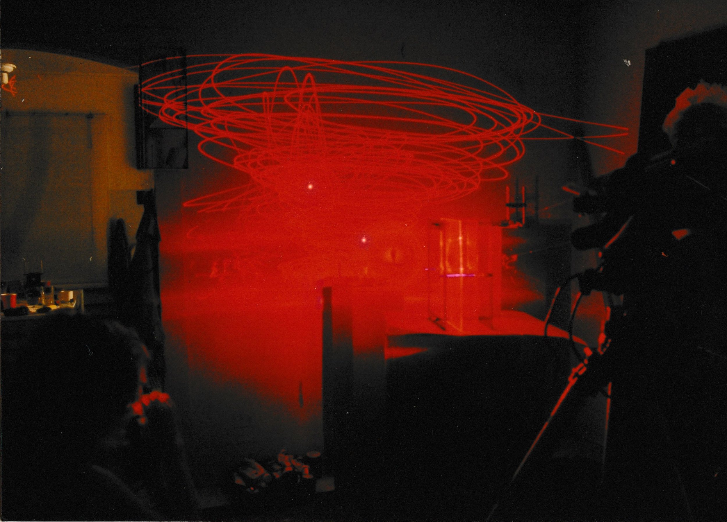 Three figures illuminated by red light stand before a gridded backdrop, surrounded by glowing laser trails.
