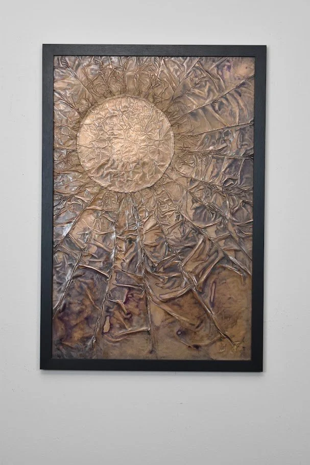 A luminous orb radiates through a fractured bronze field. Displayed framed on a white wall.