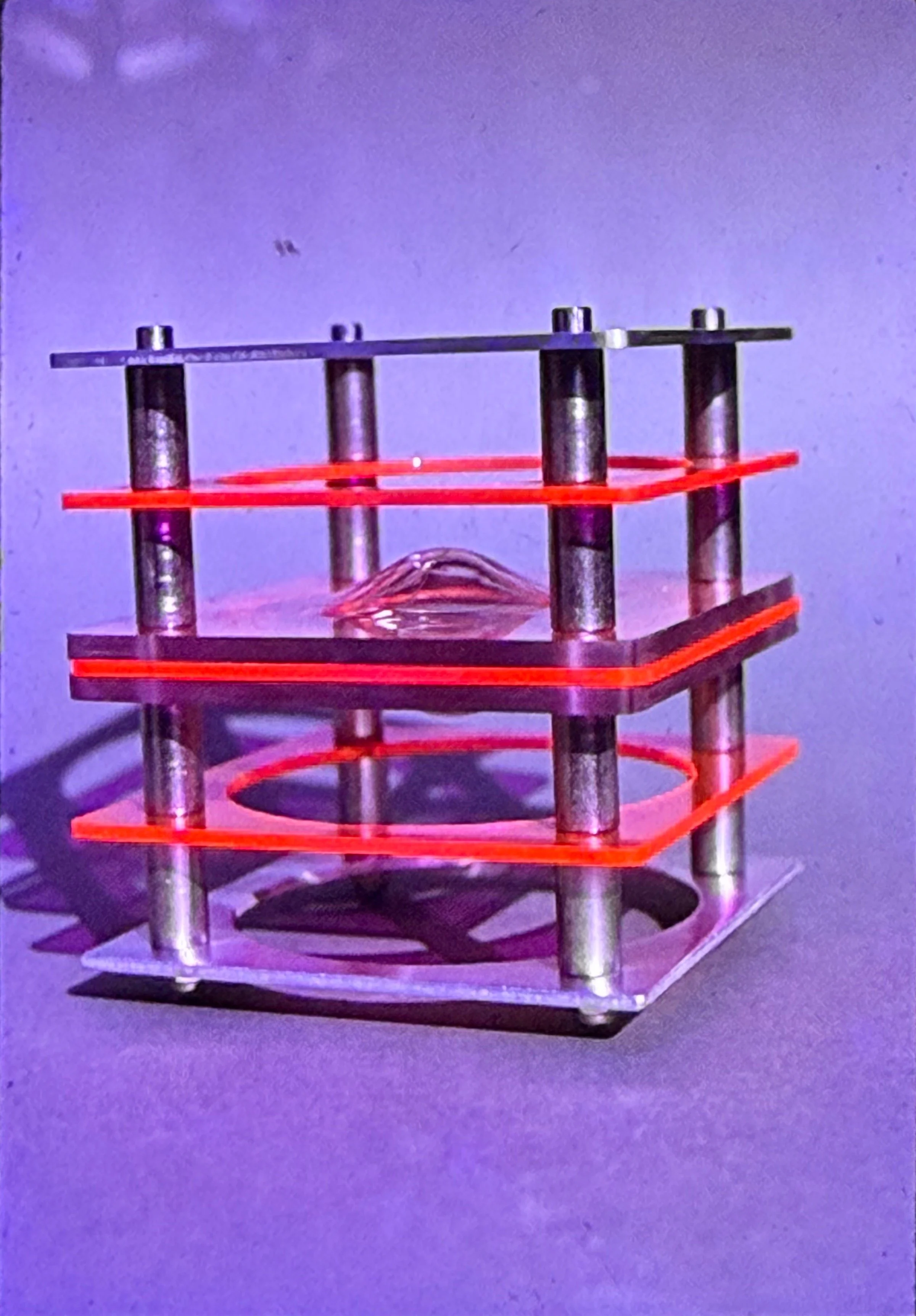 Transparent acrylic sculpture composed of stacked square planes illuminated by red linear elements and supported by vertical metal rods.