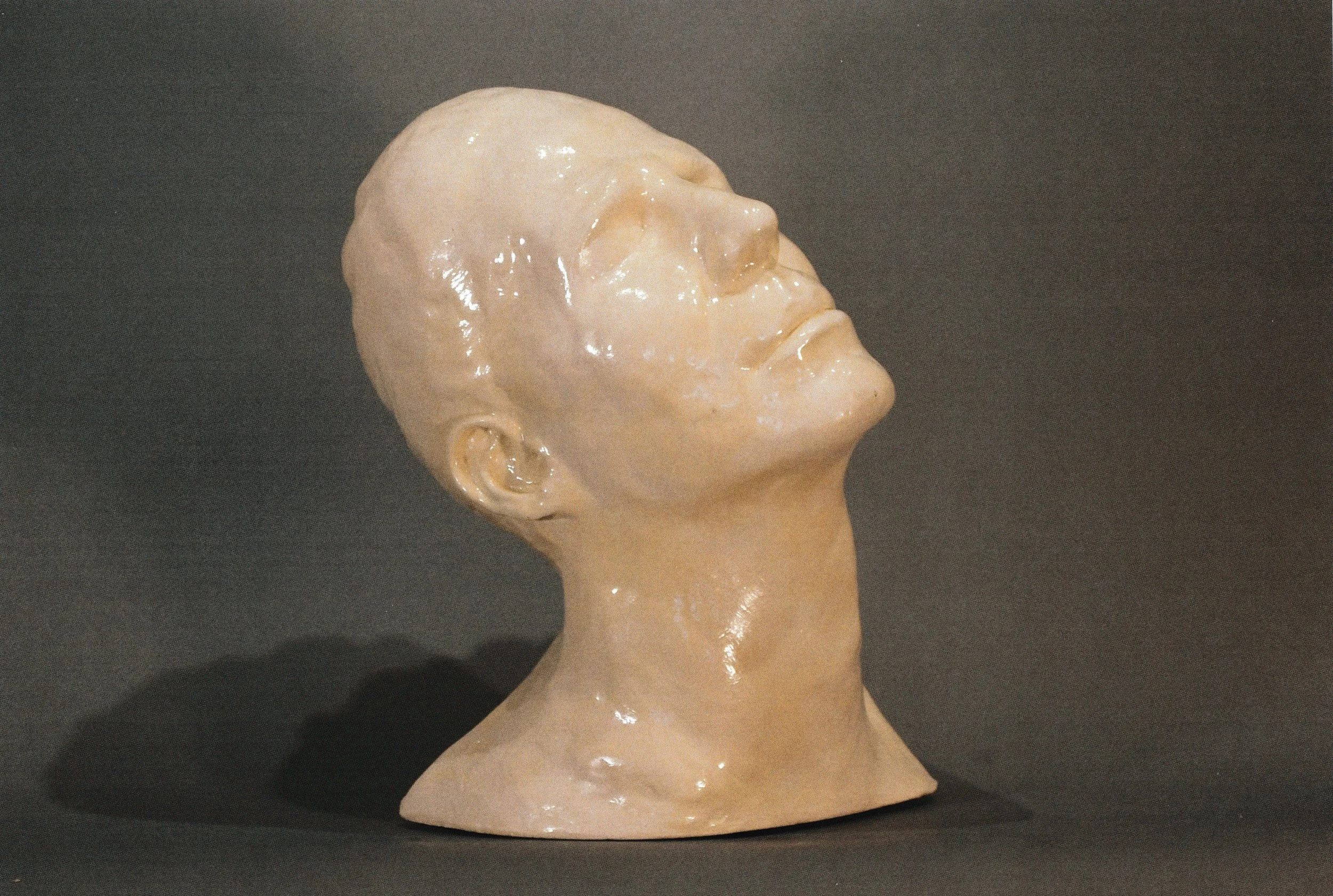 Glossy ceramic life mask portrait of a bald figure gazing skyward, illuminated against a dark backdrop.