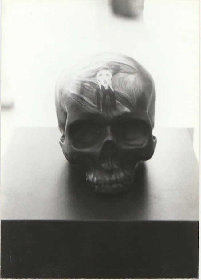 A black and white close-up view of a skull sculpture resting on a pedestal, shown from the front with deeply recessed eye sockets and a smooth, rounded cranium. The forehead contains a re-created "Scream" painting, by Edward Munch. 