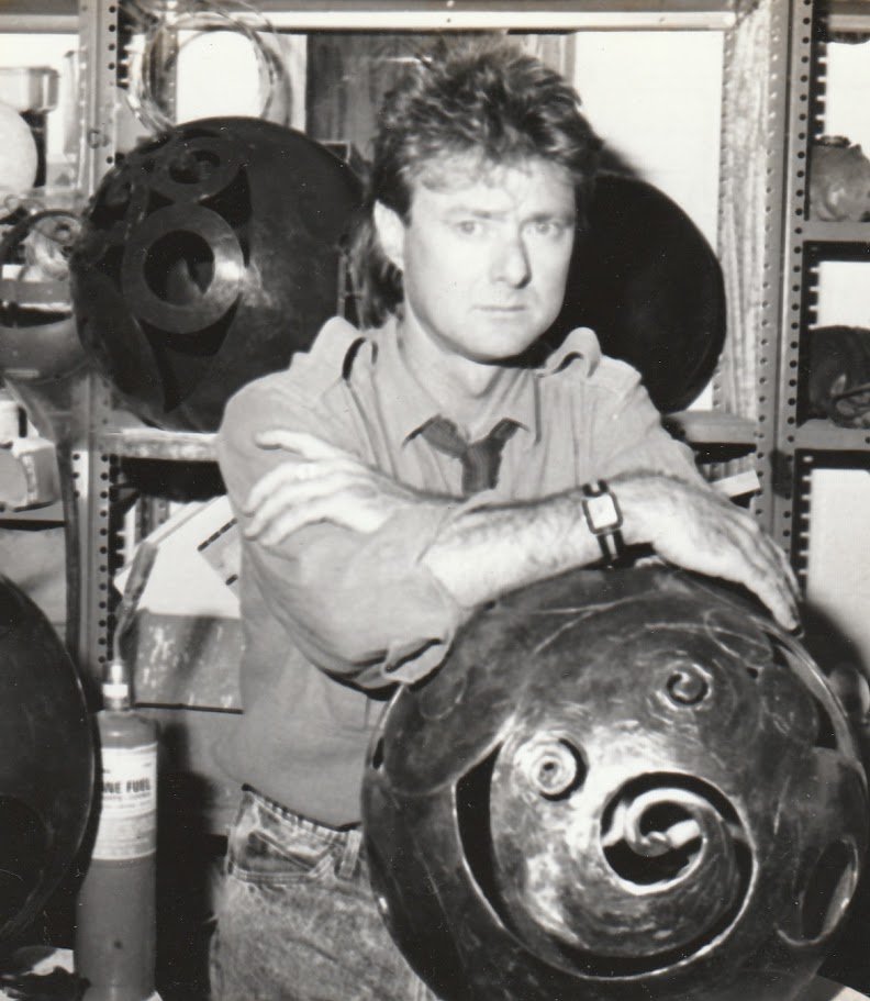 Whitney Wolf II leaning against a bronze sphere sculpture he created, in a warehouse room surrounded by other large bronze spheres. Est 1980s