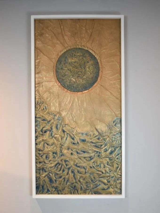 A dark orb radiates above a textured golden field. Displayed framed on a white wall.