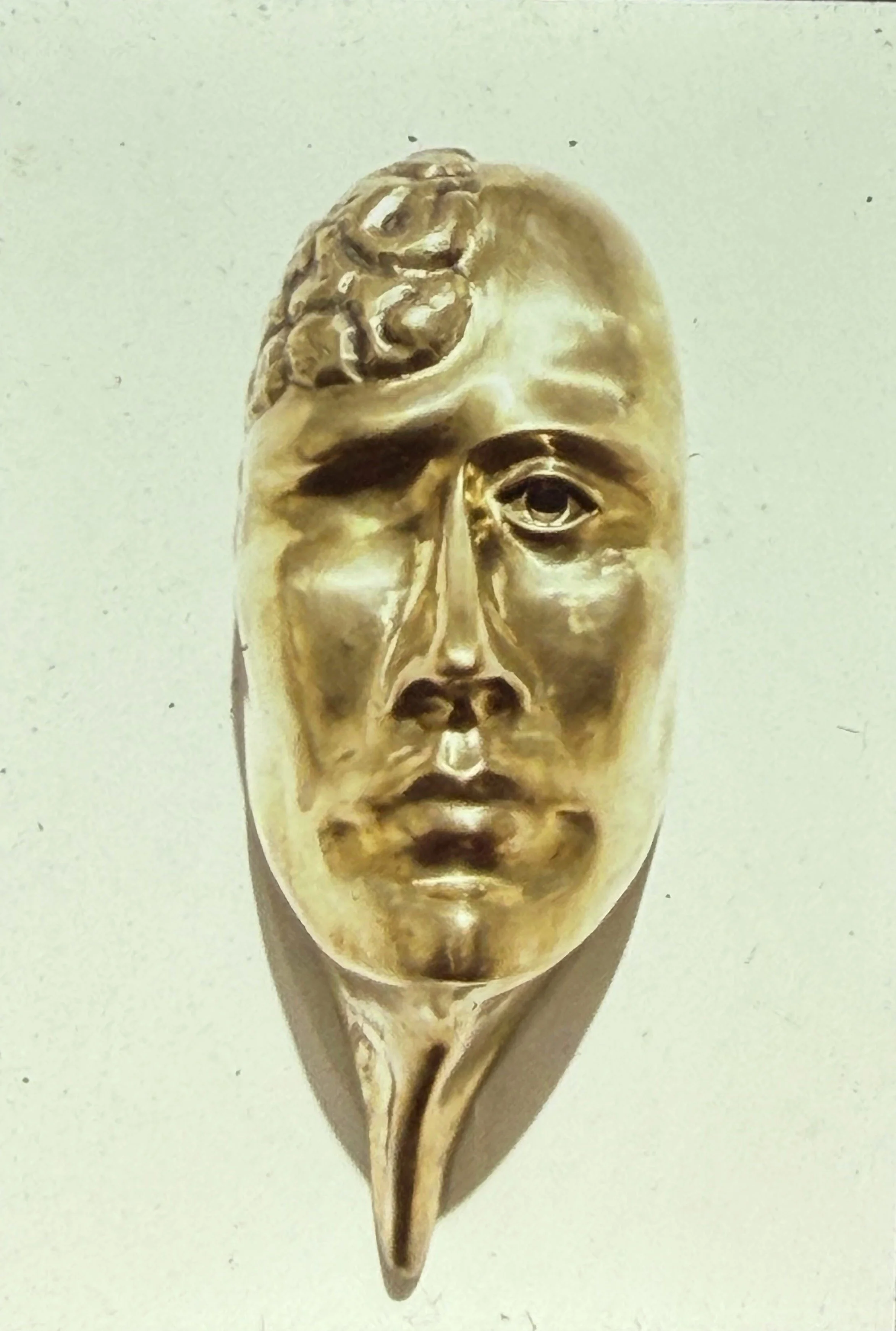 Gold-toned stylized face with smooth features and an elongated chin tapering into a pointed base. Upper left quadrant of the head has exposed brain texture.