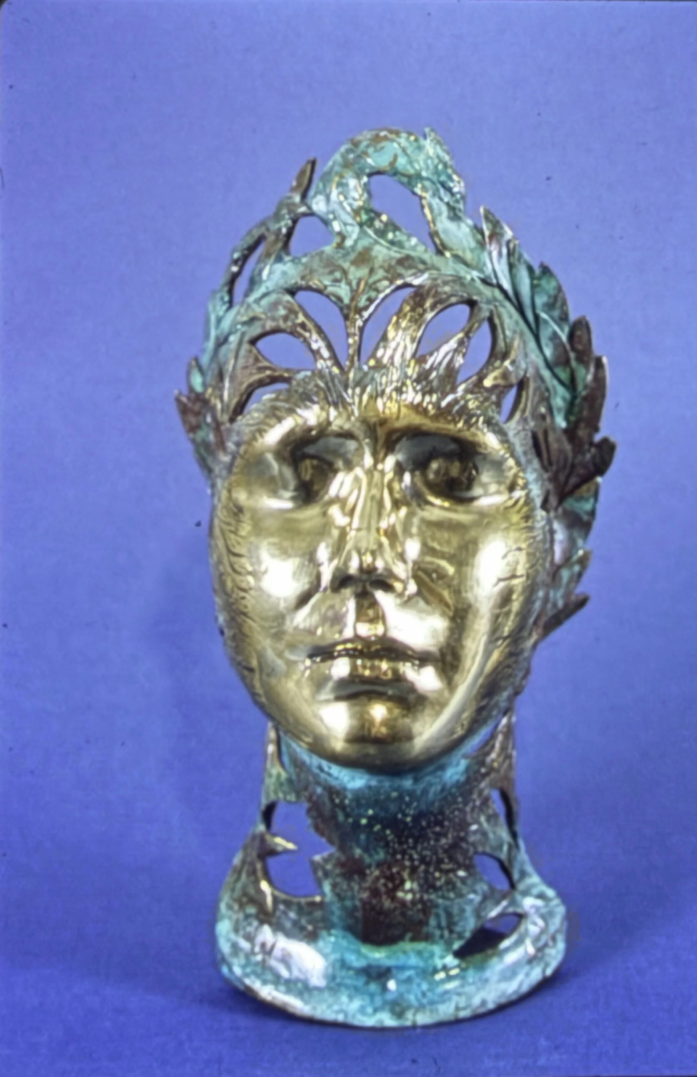 Decorative bronze face framed by openwork leaf-like forms and green patina accents resembling a crown or mask.