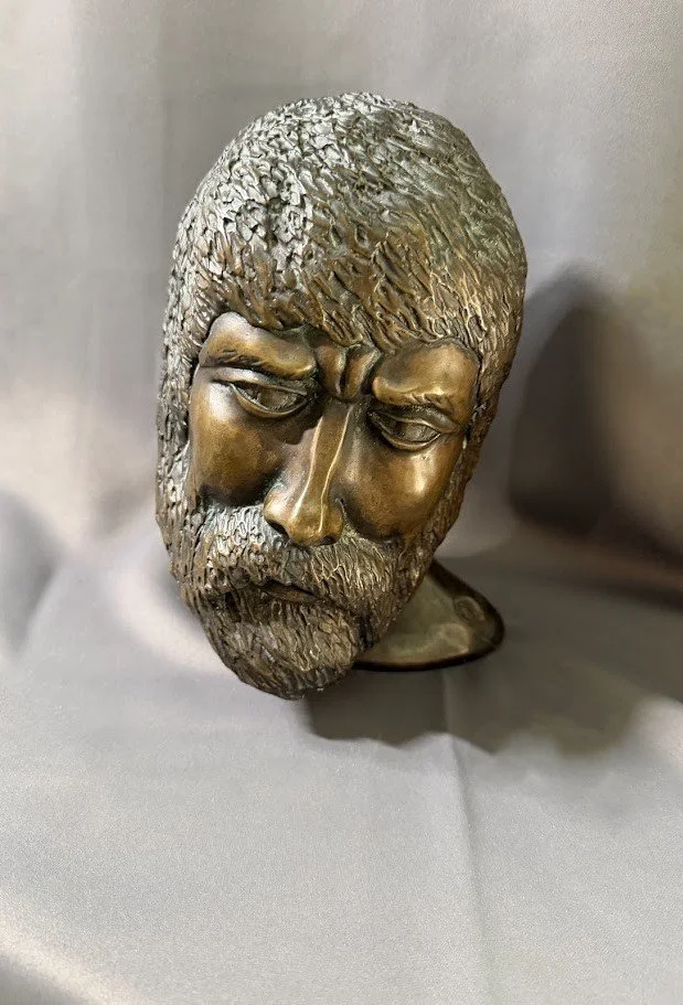 Bronze portrait head of a bearded man with eyes closed and head bowed, the rough-textured hair and beard contrasting with polished facial planes.