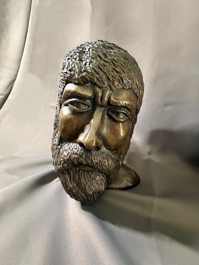 Three-quarter view of the same bronze bearded head, showing deeply carved hair, heavy brow, and contemplative downward gaze.