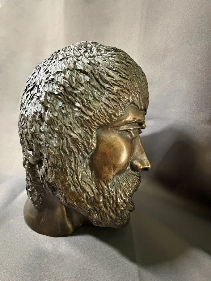 Side profile of a bronze bearded man with densely textured hair and beard, his strong nose and brow projecting sharply from the face.