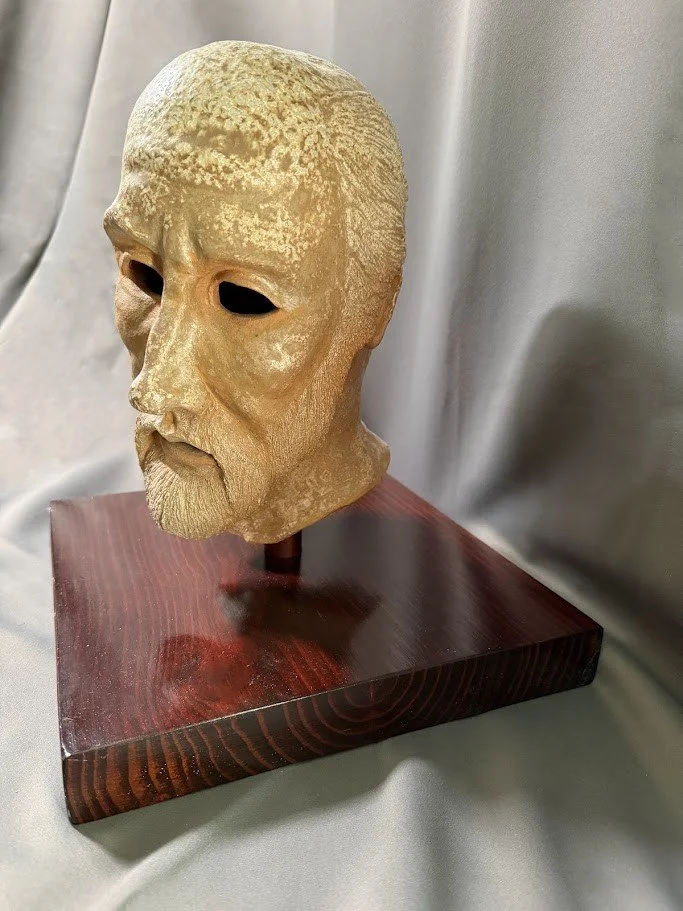 Pale mask-like sculpture of a bearded male face with dark hollow eye openings mounted on a polished reddish wood base.