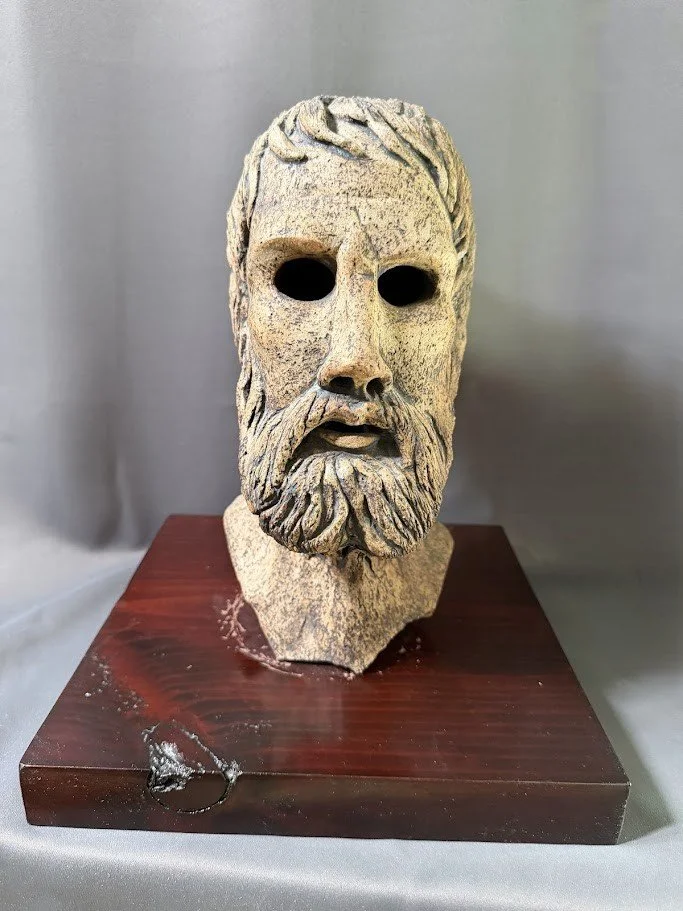 Front-facing sculpted mask with dark empty eye openings, textured hair and beard, mounted on a polished wooden base.