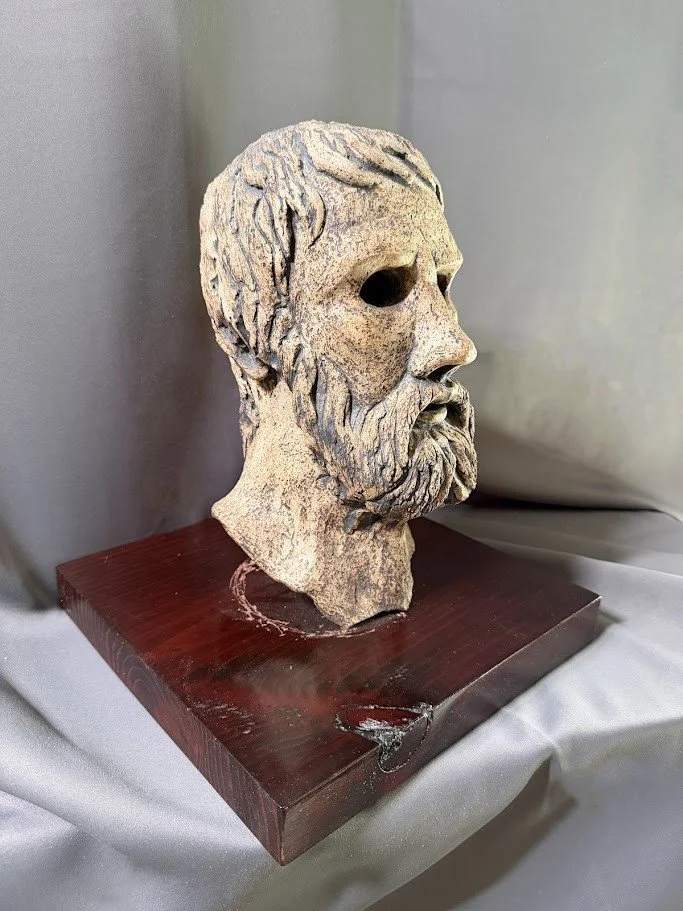 Side view of a pale mask sculpture with a long beard and deeply recessed eye sockets rising above a square wood pedestal.