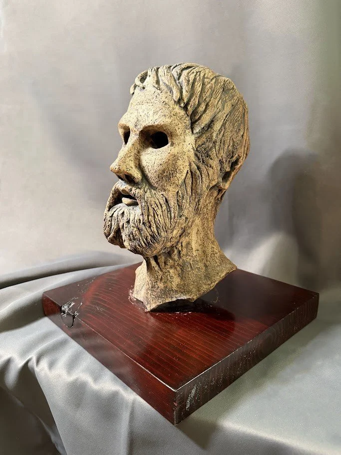 Three-quarter view of a pale classical-style mask with rough texture and dark eye cavities mounted on a wooden display base.
