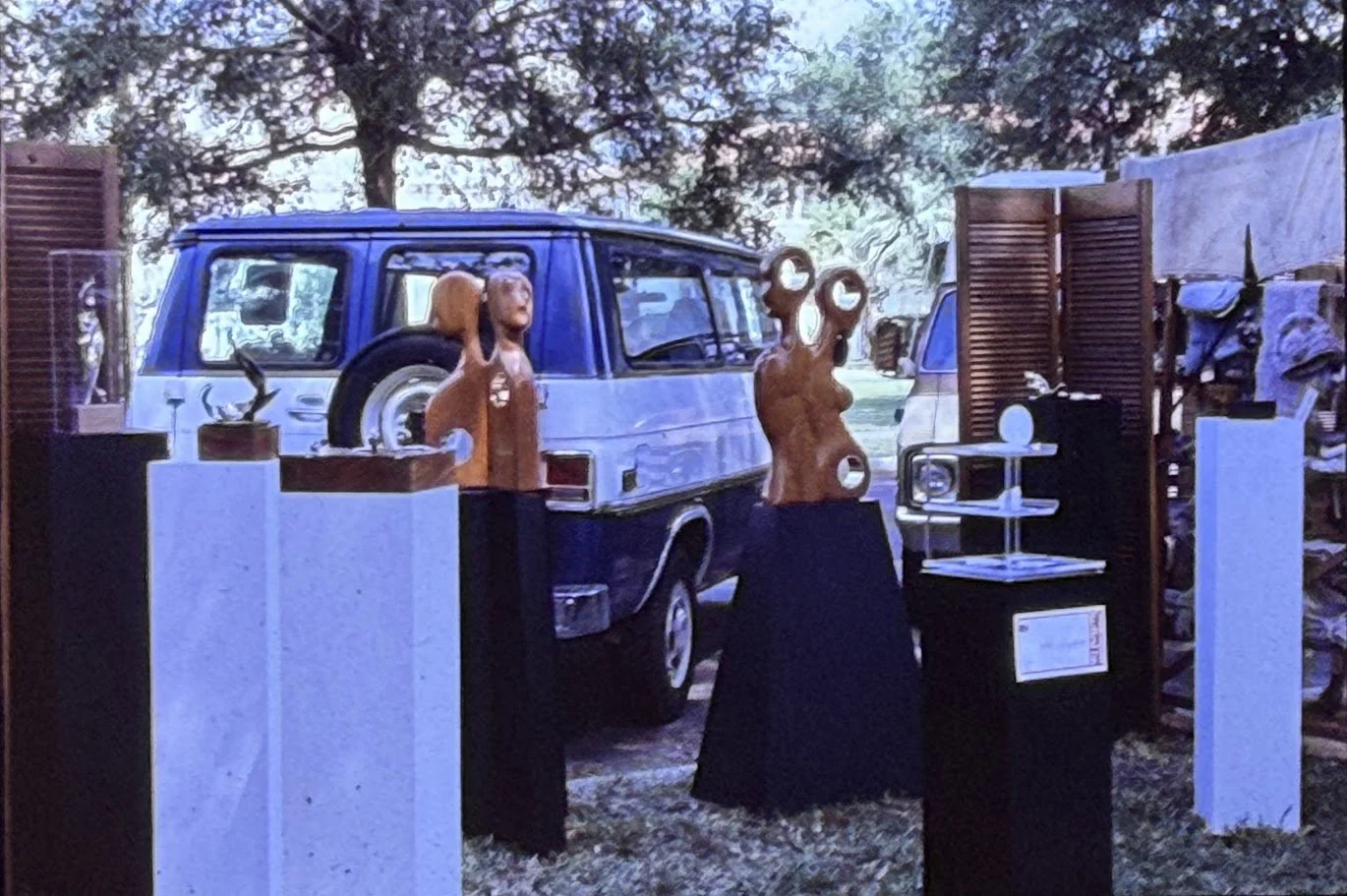 Outdoor sculpture exhibition featuring wood carvings displayed on pedestals beside a parked van.