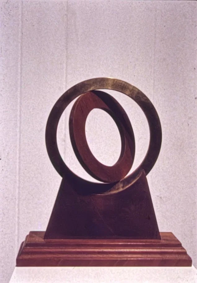 Dark stained wood sculpture formed by two intersecting oval rings mounted on a square base.
