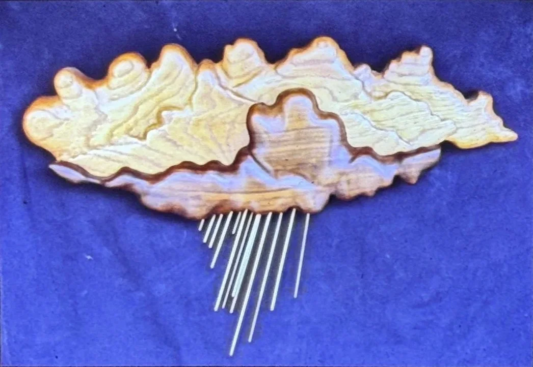 Layered wooden wall sculpture resembling clouds or a topographic landscape, with metal rods extending downward.