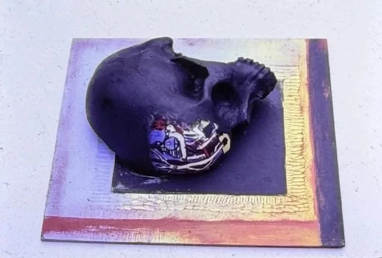 Black skull sculpture resting on a square painted base with gold, purple, and pink borders. A small colorful image is embedded along the lower side of the skull, creating a contrast between the dark surface and bright decorative detail.