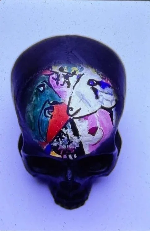 A full color close-up view of a skull sculpture resting on a pedestal, shown from the front with deeply recessed eye sockets and a smooth, rounded cranium. The forehead contains a re-created Picasso painting.