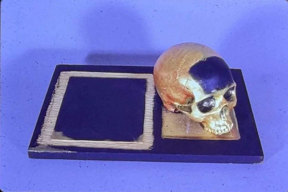 Gold and black skull sculpture resting on a rectangular black platform beside an empty square recess, photographed against a blue background.