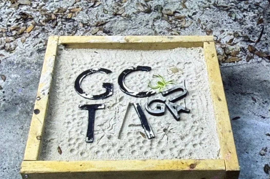 Relief panel featuring raised letters and symbols embedded in a textured square surface framed by a wooden border, further away.