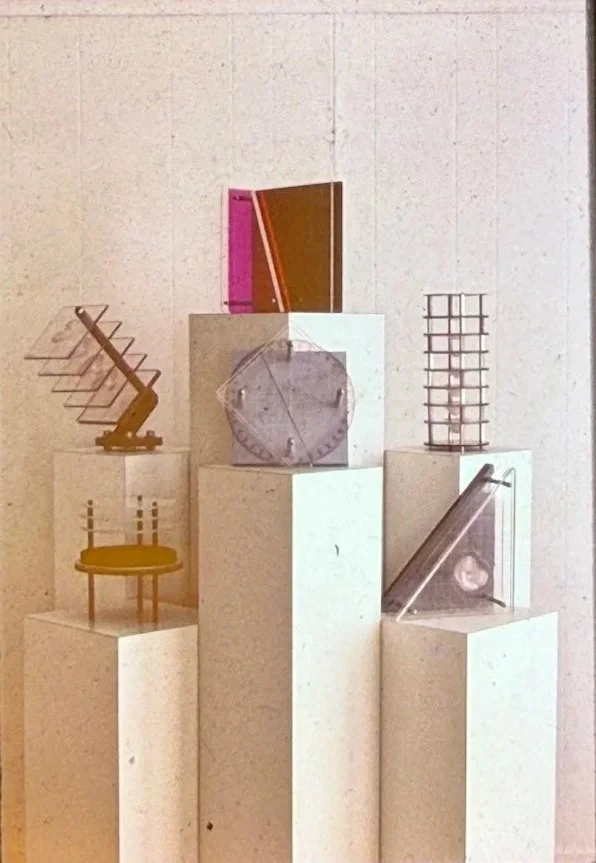 Geometric sculpture installation displayed on white pedestals, featuring abstract forms constructed from metal, acrylic, and wire elements.