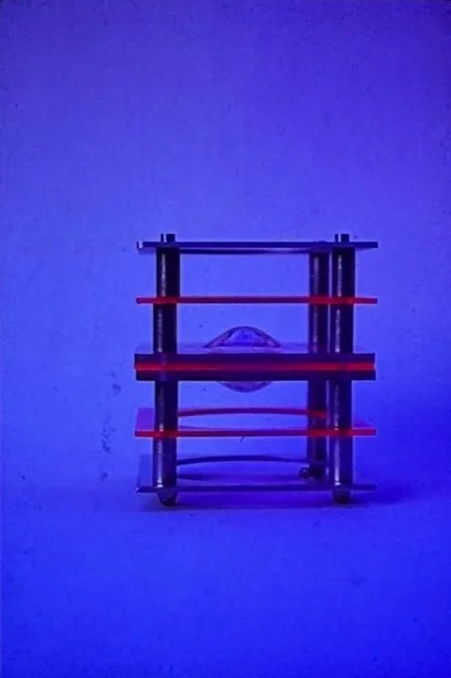 mall geometric sculpture illuminated against a blue background, featuring stacked transparent planes and red structural elements.