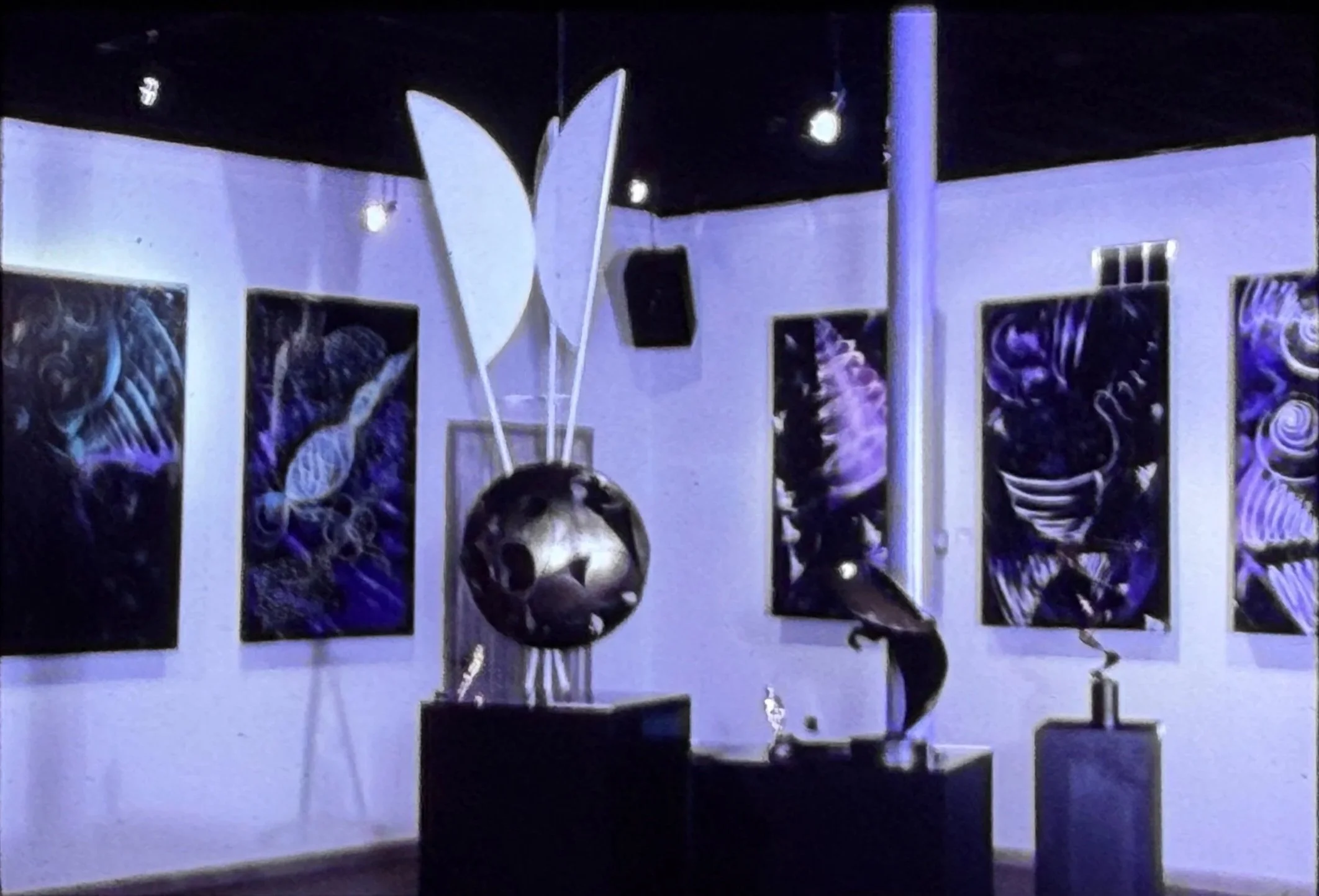 Gallery interior displaying purple-toned abstract paintings alongside freestanding sculptures.