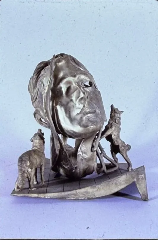 Bronze sculpture featuring a large human head emerging from a curved base, surrounded by small human and animal figures arranged around the composition. The face gazes upward while miniature figures stand beside the head and along the platform below.