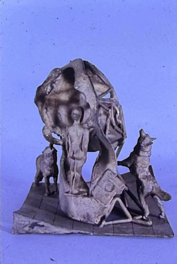 Complex bronze sculpture combining a large human head, standing figures, animal forms, and an open circular framework. Several small figures interact around the base, creating the appearance of a symbolic narrative scene.