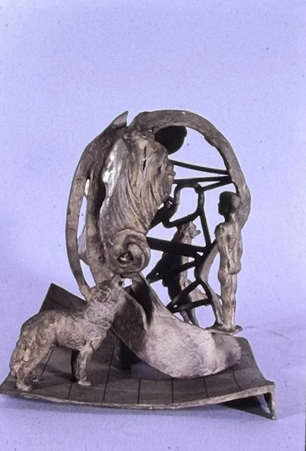 Bronze composition centered on a circular ring structure containing a human face and abstract geometric forms. Animal figures stand around the perimeter while draped and curved elements connect the various components.