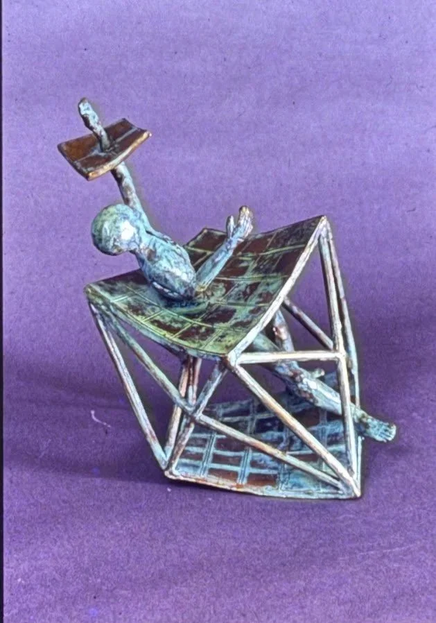 Patinated bronze figure reclining inside an open geometric framework constructed from intersecting triangular forms. The figure appears suspended within the angular structure.