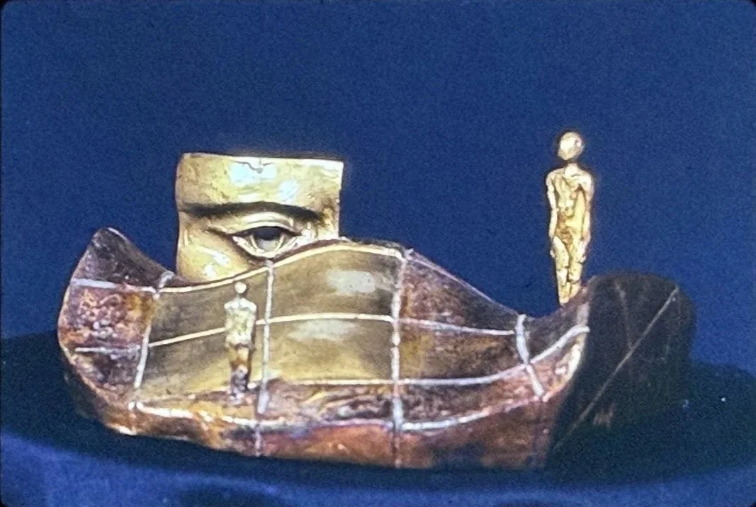 Narrative bronze sculpture featuring a large stylized eye emerging above curved architectural forms. Small standing figures occupy different levels of the composition, creating a miniature landscape-like scene.