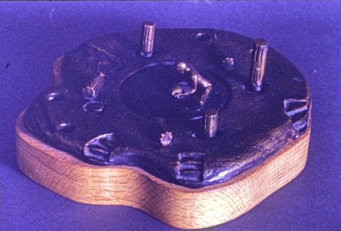 Circular bronze relief sculpture resembling a miniature environment. Tiny human figures, columns, pathways, and abstract structures surround a central opening on an irregularly shaped platform.