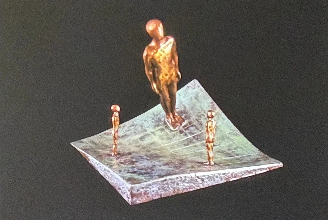 Bronze sculpture of a standing figure elevated on a sloping platform. Three smaller figures stand below, creating a sense of scale and hierarchy within the composition.