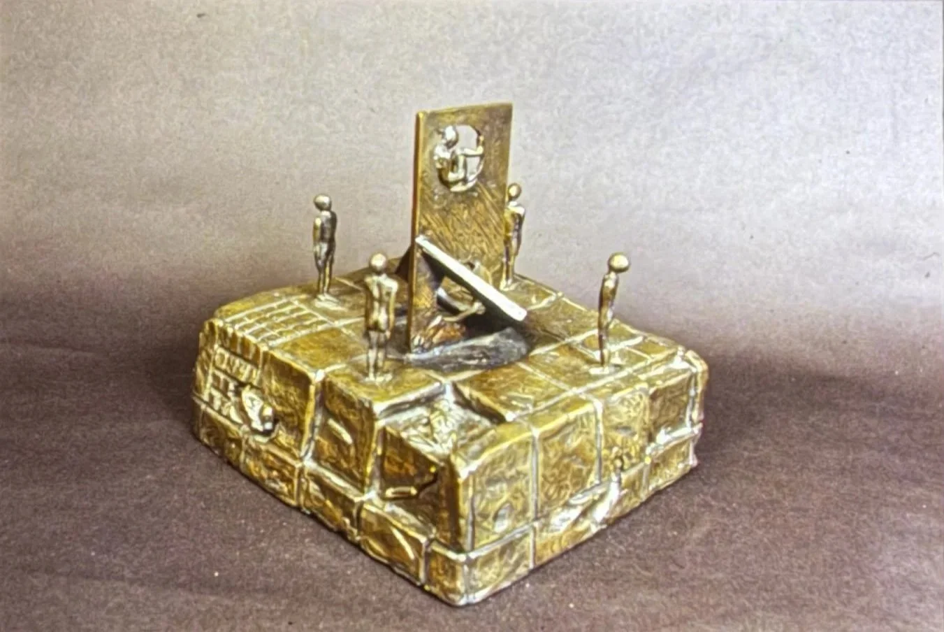 Bronze architectural sculpture featuring a raised central panel surrounded by four standing figures positioned at the corners of a rectangular platform textured to resemble stone blocks.