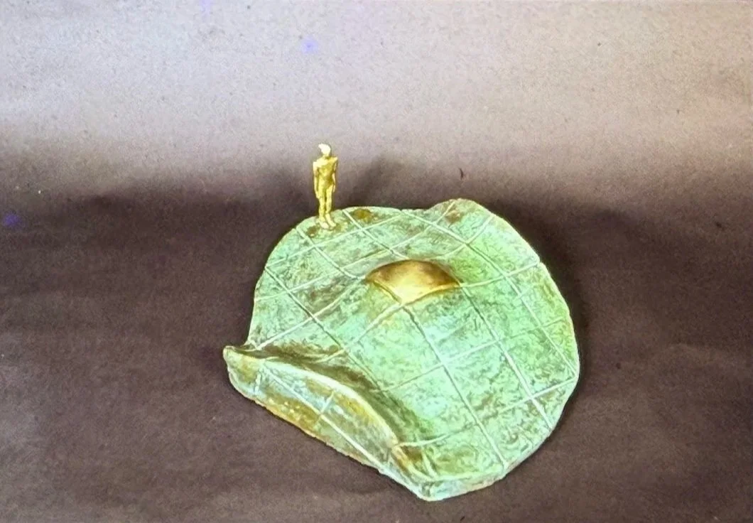 Green-patinated sculptural form resembling a curved section of landscape or shell. A miniature standing figure is positioned on top near a gold-toned recessed opening.