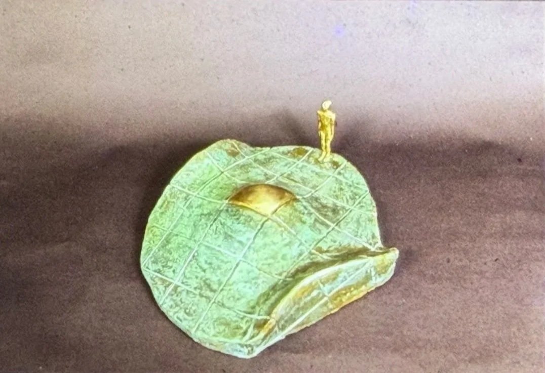 Curved green-patinated sculptural form marked by a grid pattern. A tiny standing figure appears on the crest of the curved surface near a recessed circular opening.