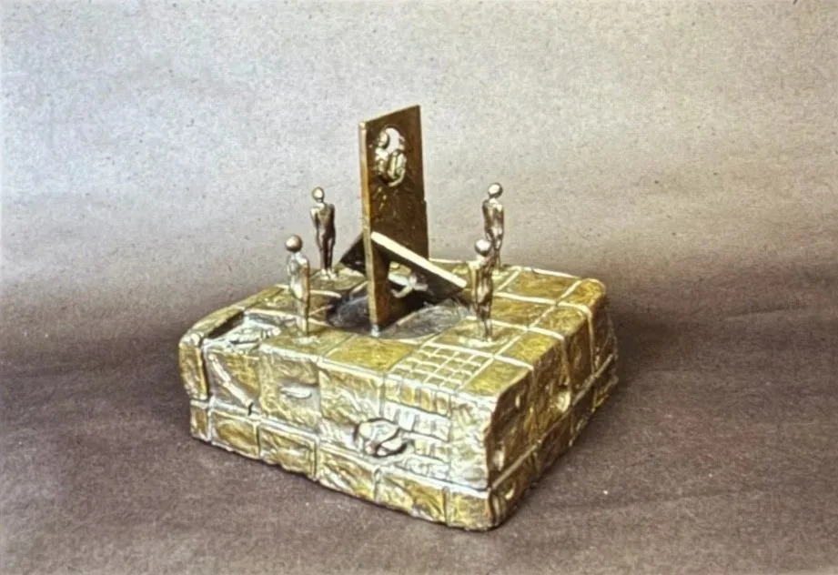 Bronze architectural scene featuring small standing figures arranged around a central panel mounted on a raised platform textured like stone blocks.