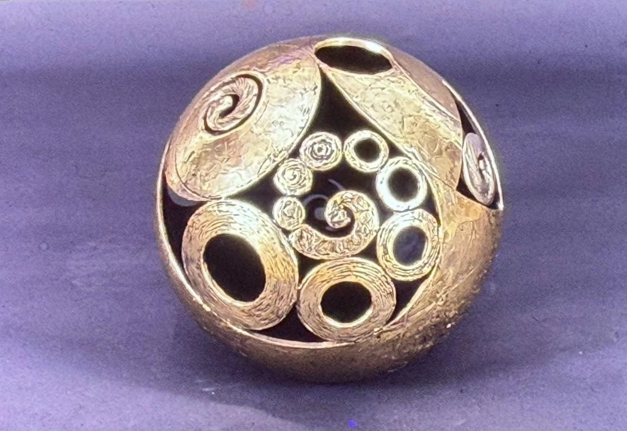 Spherical bronze sculpture decorated with spiral reliefs, circular openings, and carved patterns. The openwork surface reveals hollow spaces within the orb.