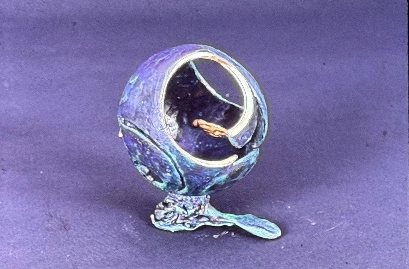 Patinated spherical sculpture resting on an organic base. A large circular opening reveals a tiny figure seated or reclining within the interior chamber.