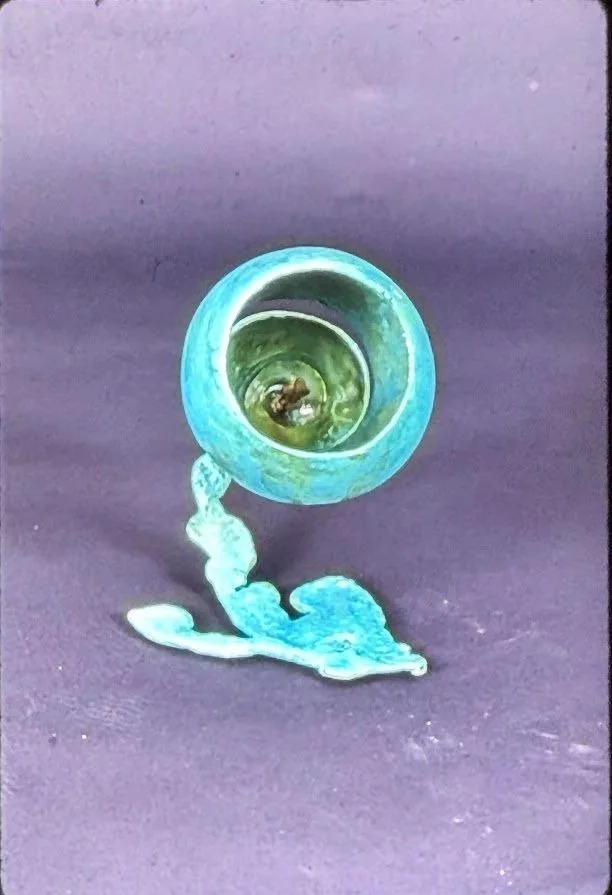 Small vessel-like sculpture with a circular opening and flowing organic base. The interior contains a tiny sculptural element visible through the opening.
