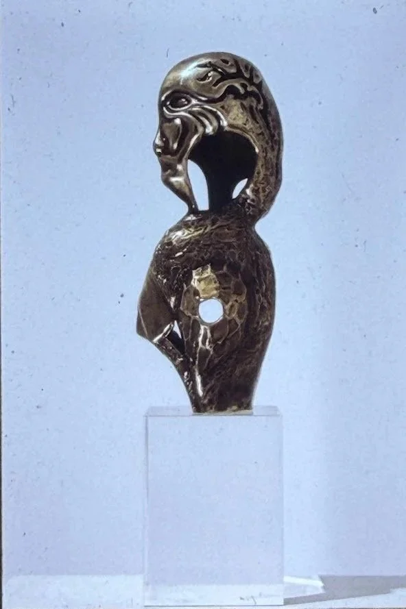 Profile view of a stylized bronze figure with a large open mouth and elongated body. The sculpture balances atop a transparent pedestal.