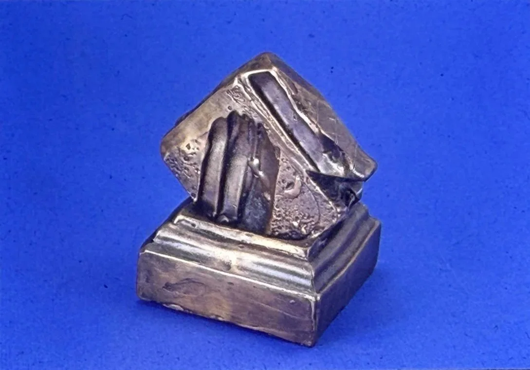 Geometric bronze sculpture composed of folded triangular planes emerging from a square plinth. The angular forms create deep shadows and sharp edges.
