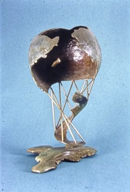 Bronze globe suspended above an open geometric framework of intersecting rods. The spherical form appears balanced above a textured organic base.