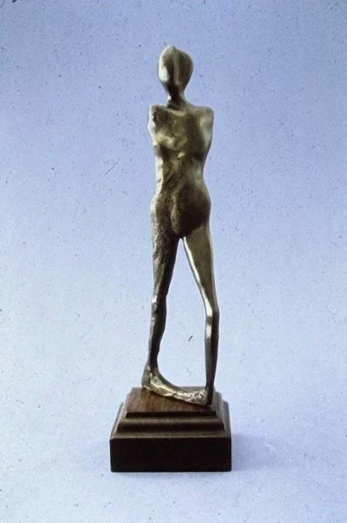 Slender bronze female figure standing upright on a stepped pedestal, with elongated limbs and simplified anatomical features.