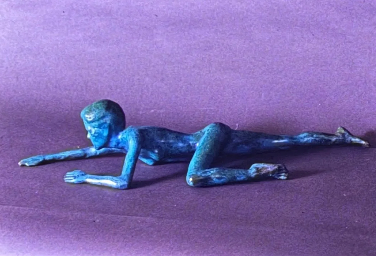 Blue-green patinated bronze figure crawling low to the ground, stretched forward with one arm extended and legs trailing behind.