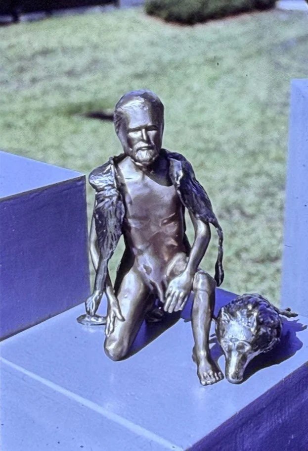 Bronze wolf-headed seated figure displayed outdoors, combining human anatomy with animal symbolism.