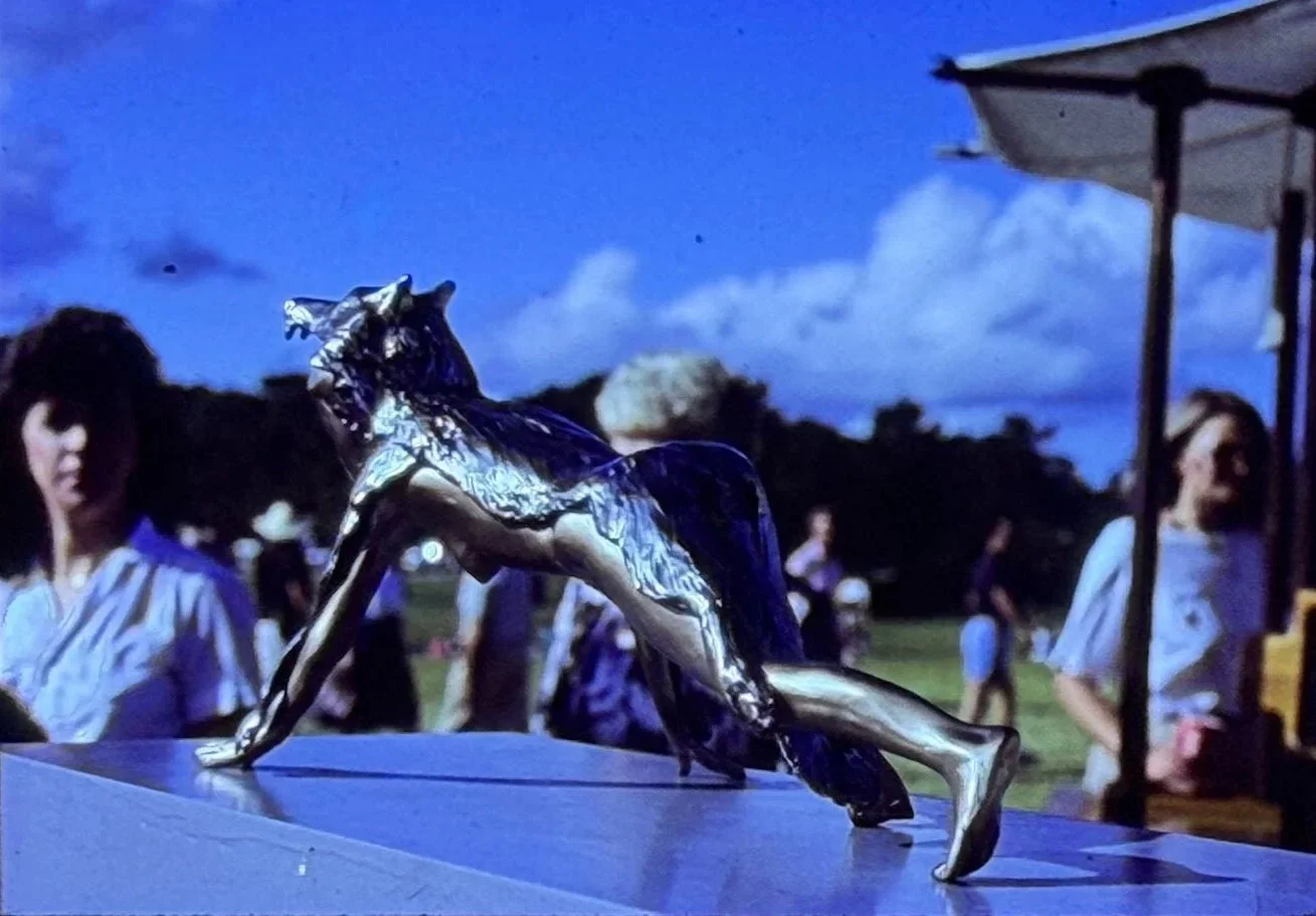 Bronze wolf-like creature lunging forward with elongated limbs and an alert posture, displayed outdoors during an exhibition.