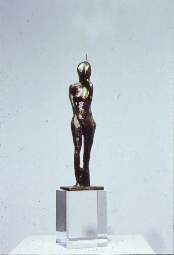 Slender bronze sculpture of a standing female figure with elongated proportions, arms held behind her back, and one leg crossed in front of the other, mounted on a clear pedestal.