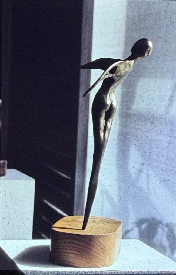 Slender abstract figure leaning dramatically backward from a wooden base, balanced on elongated legs.