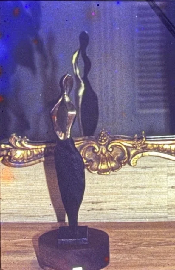 Slender abstract bronze figure with a twisting silhouette, reflected in an ornate mirror. The elongated body narrows into a dark pedestal, emphasizing elegance and vertical movement.